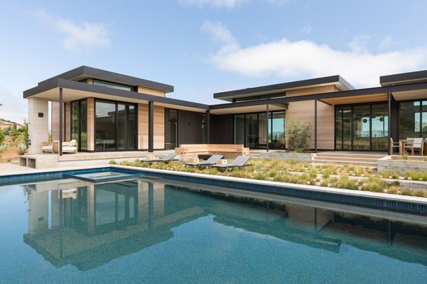 A low-slung contemporary residence arranged around a landscaped pool courtyard. The home features broad flat roof planes, dark horizontal fascia, warm timber-toned wall cladding, extensive floor-to-ceiling glazing, and sheltered outdoor living areas. A large swimming pool dominates the foreground, with a paved terrace, timber bench seating, lounge chairs, and drought-tolerant planting around the building.