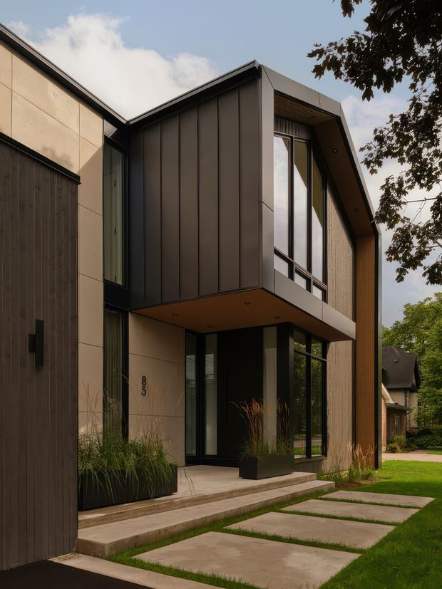 Contemporary multi-level residential facade viewed from the front, featuring dark vertical metal cladding, pale large-format wall panels, extensive black-framed glazing, a projecting upper floor, and a deep wood-lined roof overhang. Concrete steps and pavers lead to the recessed entry, with integrated planter boxes and landscaped lawn. The image also shows neighbouring residential context and mature trees.