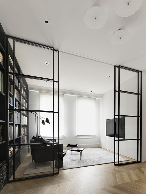 Bright, refined living room with a neutral palette and herringbone timber flooring. The room features full-height black metal-framed glazed partitions and doors, a large built-in black bookcase, a dark sofa and armchairs, low round coffee tables, a pale area rug, and a wall-mounted television concealed within white built-in joinery. High windows, decorative ceiling cornice, and sculptural circular ceiling lights add architectural detail.