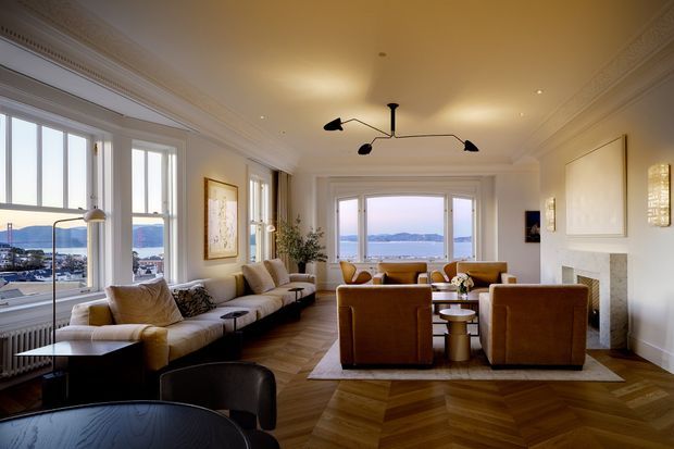 Elegant waterfront living room with tall windows overlooking a bay, a central seating arrangement, white walls, ornate ceiling cornices, a marble fireplace, and warm herringbone timber flooring. The room combines contemporary furniture with refined traditional architectural detailing and warm layered lighting.