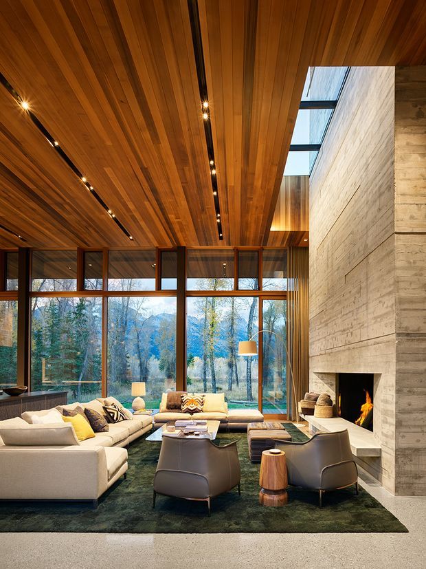 A spacious, warm living room with floor-to-ceiling timber-framed glazing overlooking a wooded landscape. The room features a dramatic full-height concrete or textured masonry fireplace, a richly timber-lined ceiling with integrated linear spotlights, large cream sectional sofas, dark leather lounge chairs, a low coffee table, and a dark green rug. A clerestory-like skylight or glazed roof opening brings additional daylight into the space.