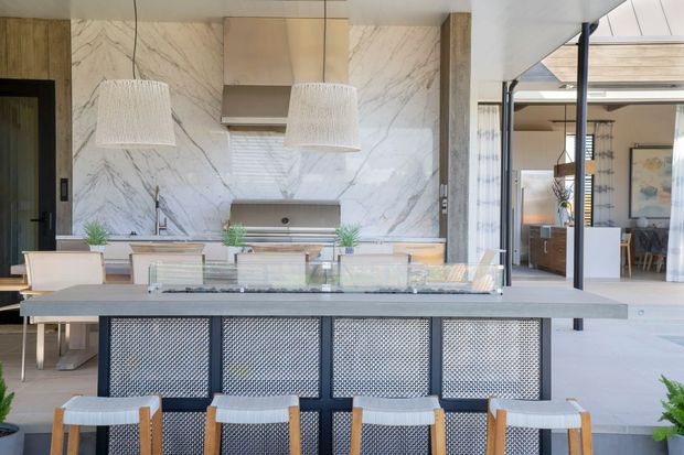 A contemporary open-plan kitchen and dining area centered on a large island with a dark metal mesh base, pale stone countertop, and several timber-framed stools. Behind it is a full-height veined marble backsplash, streamlined cabinetry, integrated appliances, and a sink beneath a concealed or minimal window treatment. Two oversized textured pendant lights hang above the island. Glazed openings connect the kitchen to a bright dining and living area, with a black-framed internal window or screen adding contrast.