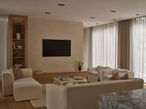 A refined contemporary living room with warm neutral tones, built-in timber shelving and low cabinetry, a wall-mounted television, cream upholstered seating, a pale coffee table, and full-height sheer and draped curtains across expansive glazing. A marble-topped surface is visible in the foreground, and recessed ceiling lighting creates a calm, softly lit atmosphere.