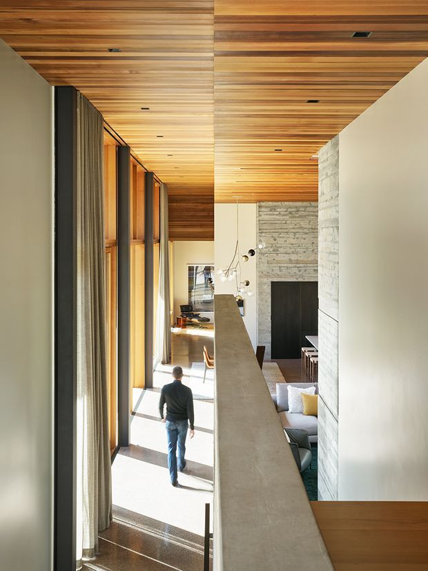 A tall, light-filled contemporary interior viewed from an elevated level, featuring a long open void, floor-to-ceiling glazing with curtains on the left, a warm slatted timber ceiling, exposed pale masonry walls, and a concrete balustrade overlooking a living area with a fireplace and furnishings below. A person provides scale.