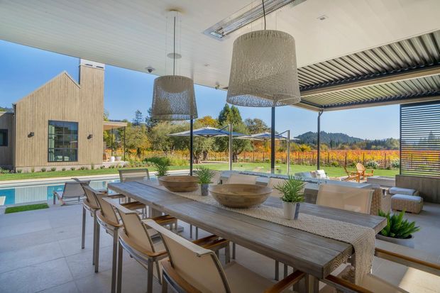 A spacious, contemporary covered outdoor dining and entertaining area beside a swimming pool. A long timber dining table with upholstered chairs sits beneath large woven pendant lights, with an outdoor kitchen or bar zone to the right. Open slatted screening and slim structural posts frame views toward the landscaped property and a modern gabled building beyond.
