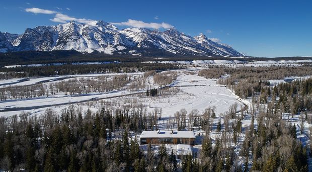 Aerial exterior view of a snow-covered rural or alpine landscape with a long, low building nestled among dense trees in the foreground. A dramatic snow-capped mountain range dominates the background, with open fields, winding tracks, and scattered woodland visible across the valley.