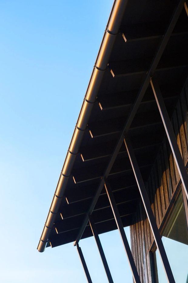 Upward-looking close-up of a building exterior showing a deep dark roof eave, exposed structural supports, a metal gutter and downpipe, and vertical timber wall cladding beside glazing. The strong diagonal composition emphasizes the roof edge and external structure against a clear blue sky.
