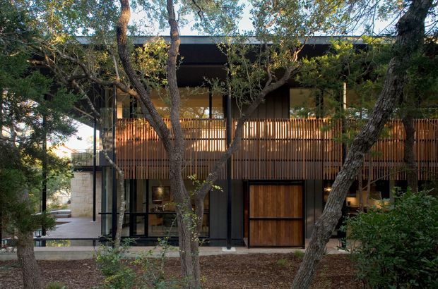 A two-storey contemporary residential facade nestled among mature trees. The building features dark vertical structural elements, extensive glass, warm timber slat screening and balcony railings, a timber garage or entry door, and a broad flat roofline. The wooded landscaping partially obscures the architecture.