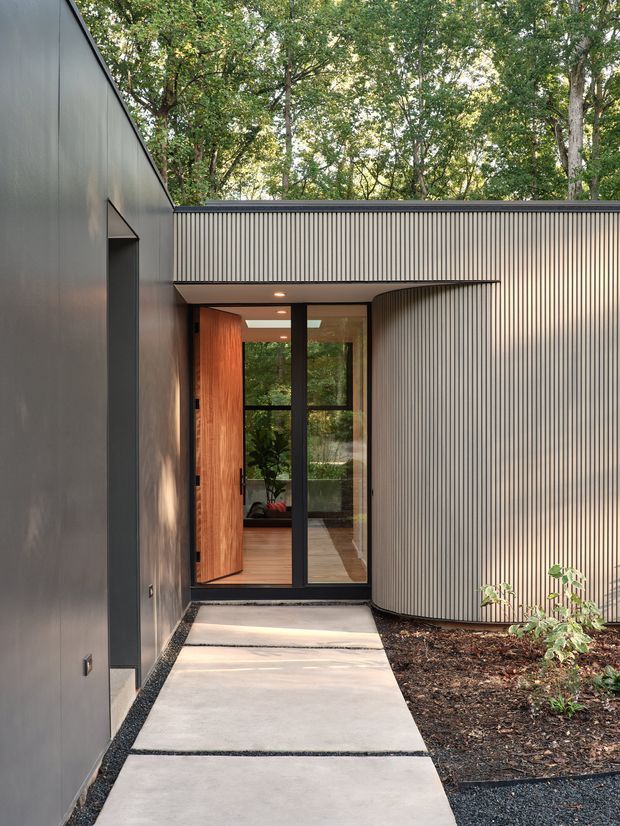 A modern residential entrance viewed from an exterior walkway. The composition features dark smooth wall cladding on the left, a curved facade clad in narrow vertical ribbed panels on the right, and a glazed entry with a warm timber pivot door and black-framed glazing. Concrete stepping slabs lead through landscaped planting toward the doorway, with mature trees visible beyond.