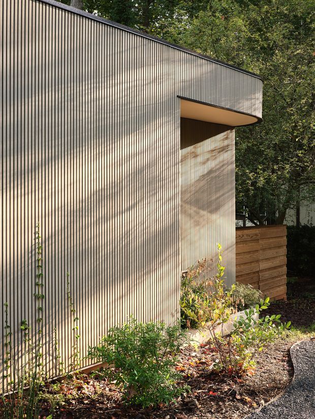 Exterior view of a compact contemporary building set within a wooded landscaped site. The facade is clad in closely spaced vertical ribbed boards with a warm grey-beige finish, and features a projecting curved-edged canopy or opening with a dark underside. A timber retaining wall, planted garden bed, and asphalt path occupy the foreground, while mature trees surround the structure.