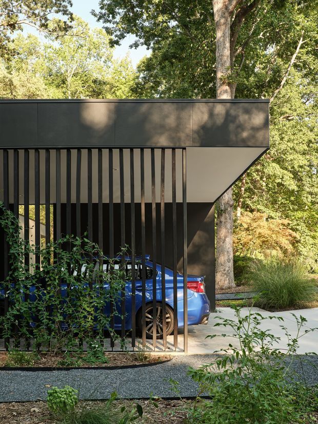 Exterior architectural photograph of a contemporary carport integrated into a wooded landscape. The structure has a flat roof with dark metal cladding, a sharply angled soffit, and a screened side formed by closely spaced vertical dark slats. A blue car is parked beneath the canopy, with a concrete driveway and landscaped garden surrounding it.