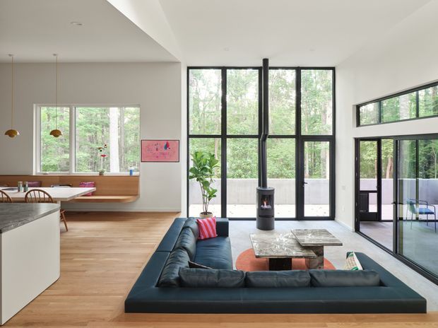 Open-plan contemporary living and kitchen interior with a dark blue sectional sofa facing a low coffee table and freestanding black wood stove. The kitchen includes a pale island, timber flooring, built-in window seating and pendant lights. Large black-framed floor-to-ceiling glazing opens toward a leafy outdoor setting, with a high horizontal clerestory-style window along the right wall. The space is bright, refined and connected to nature.