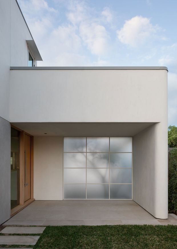 A minimalist modern residential facade viewed from the front, featuring smooth white rendered walls, a projecting upper volume, and a recessed covered entry terrace. The terrace includes a warm timber-framed glazed door on the left and a large translucent grid-patterned glass panel at the rear. A rounded wall corner, concrete paving, stepping stones, and a narrow strip of lawn complete the composition. The image has a clean, quiet architectural character with soft natural daylight.