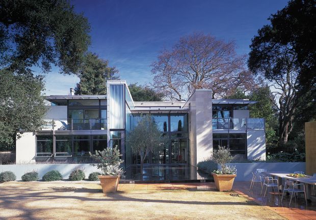 A low-rise contemporary residential building viewed from the landscaped front courtyard. The symmetrical facade combines white rendered walls, extensive floor-to-ceiling glazing, dark metal-framed windows, and a projecting flat roof. A tall glazed vertical element marks the central entry, with outdoor dining furniture and large planters in the foreground, surrounded by mature trees.