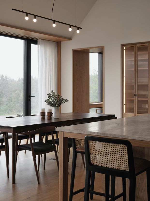 A warm, refined dining and kitchen interior with a dark timber dining table and chairs beside a large stone-topped island. Floor-to-ceiling glazing and sheer curtains bring in soft natural light and views of trees. The room features timber-framed openings, built-in glazed timber cabinetry, timber flooring, and a linear black track light.