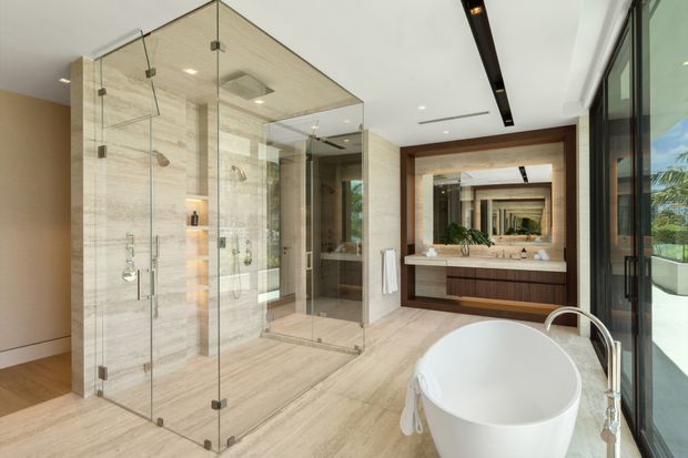 Spacious contemporary bathroom with a freestanding white oval bathtub beside full-height glazing, two large frameless glass shower enclosures, a double vanity with dark wood cabinetry and a pale stone benchtop, and large light beige stone-look floor and wall surfaces. Recessed lighting and a dark linear ceiling feature add contrast, while the mirror reflects the vanity and shower area.