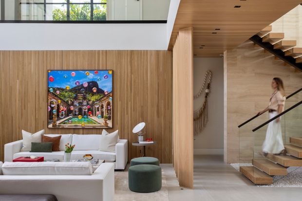 A refined contemporary living room opening to a stair hall. The room features a white sectional sofa, low white coffee table, dark green ottomans, a vertically slatted timber feature wall with colourful artwork, pale stone or concrete-look flooring, and extensive light timber joinery. A sculptural open staircase with timber treads and black railing rises beside a pale stone-clad wall, while a corridor and upper-level glazing are visible beyond.