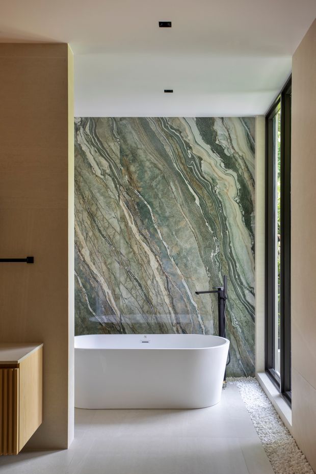 A refined contemporary bathroom centered on a freestanding white bathtub. The back wall is clad in dramatic green-grey and cream veined natural stone, contrasted with pale smooth flooring and warm timber-toned built-in cabinetry. A black wall-mounted tap and shower fitting, recessed ceiling lights, and full-height dark-framed glazing add a modern, luxurious feel. White decorative pebbles line the window edge.