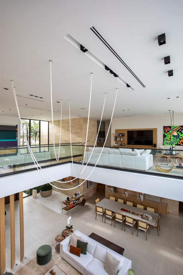 A tall, double-height contemporary residential living space viewed from an upper-level landing or mezzanine. The open-plan interior includes a white sectional sofa and television area above, a dining table below, glass balustrades, pale timber wall panelling, polished light flooring, and a dramatic sculptural pendant made from long white suspended rods. Modern recessed and linear ceiling lighting, colourful artwork, and an open void connect the two levels.