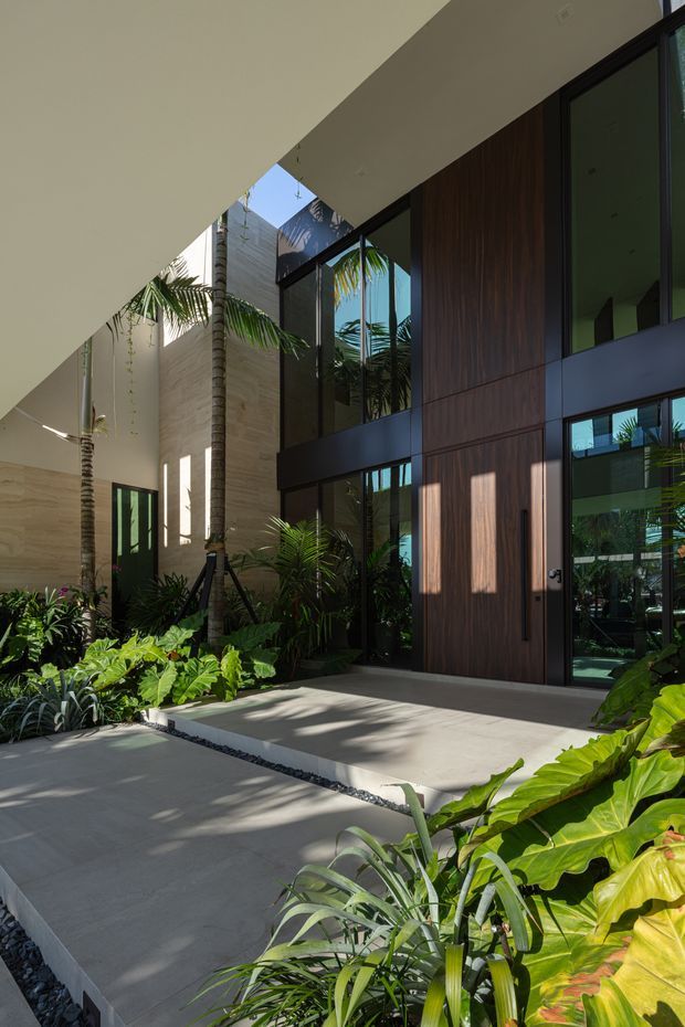 A contemporary tropical residential entrance courtyard with a tall, partially covered entry canopy, extensive floor-to-ceiling dark-framed glazing, and a large timber-clad pivot door. Lush tropical planting surrounds broad light-grey paved paths, while strong sunlight creates patterned shadows across the hardscape and facade. The space has a refined indoor-outdoor connection and a tall, airy volume.