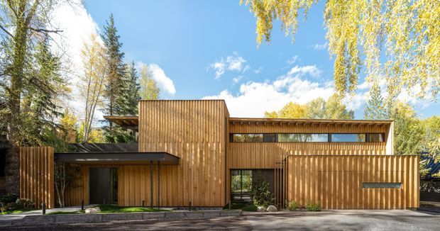 Exterior view of a contemporary multi-level residence set among mature trees. The building has a strongly horizontal composition, extensive vertical timber cladding, a low-pitched roof, deep overhangs, narrow clerestory-like glazing, and a partially enclosed carport or garage element at the right. The warm wood facade contrasts with dark window and door frames and the surrounding leafy landscape.