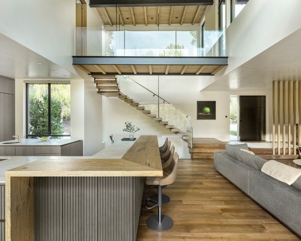 A bright, open-plan contemporary interior combining kitchen, dining, and living areas beneath a dramatic double-height void. The kitchen features a large timber island with vertically fluted dark cabinetry and bar seating, while warm timber flooring and ceiling boards continue through the space. A sculptural open-riser stair with glass balustrade leads to a mezzanine or upper landing enclosed by clear glazing. White walls, extensive glazing, a grey sectional sofa, and vertical timber screening complete the refined natural palette.