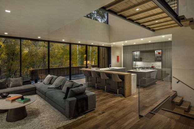 Open-plan contemporary living, dining and kitchen area with a large charcoal sectional sofa, kitchen island with bar seating, extensive floor-to-ceiling glazing overlooking trees, warm timber flooring, and a dramatic exposed timber ceiling structure above the kitchen. A stairway with glass balustrade is visible at right.