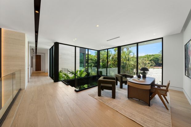 A bright, spacious contemporary interior combining a hallway and living area. Light timber flooring and built-in timber cabinetry extend along the left wall, while a low-profile seating arrangement and desk occupy the glazed corner. Full-height black-framed glazing overlooks lush greenery and water, with an internal planted lightwell or courtyard visible beyond the glass partitions. Recessed lighting, a clean white ceiling, and restrained furnishings create an airy, refined atmosphere.