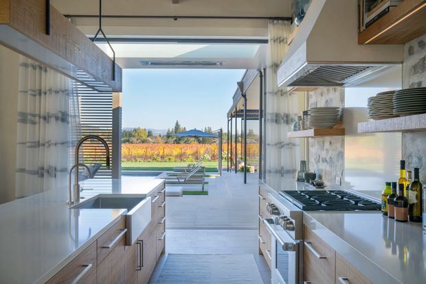 A bright, modern galley kitchen with parallel banks of light timber cabinetry, pale stone or engineered-stone benchtops, twin sinks, integrated appliances, a large gas cooktop and stainless-steel rangehood. A fully glazed opening at the end connects the kitchen to a covered outdoor area and landscaped vineyard-like garden beyond. The space has a clean, contemporary fit-out with strong indoor-outdoor connection and abundant natural light.