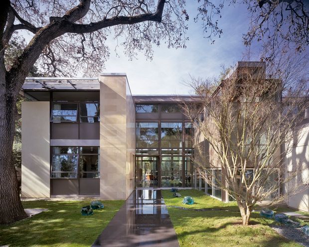 Exterior view of a contemporary multi-wing building arranged around a landscaped courtyard. The facade combines extensive dark-framed glazing, pale solid wall planes, and a projecting roof canopy, with a reflective paved walkway leading toward a glazed central entry. Mature trees and lawn frame the architecture.
