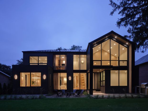 A dusk exterior photograph of a contemporary multi-level residence with a dark, rectilinear facade and a prominent steeply pitched gable-like glazed volume. Extensive floor-to-ceiling windows reveal warm interior lighting, a stairway, and double-height living space. The home sits beside a landscaped lawn with mature trees and neighbouring structures visible at the edges.