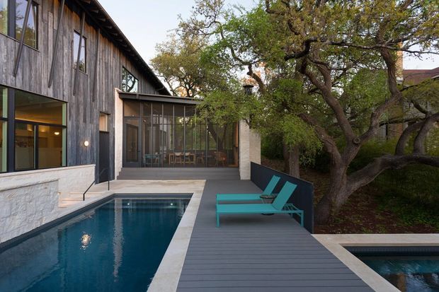 A contemporary residential courtyard viewed from an outdoor pool deck. The house features dark weathered vertical timber cladding, extensive glazing, and a low-pitched roofline. A narrow swimming pool runs alongside a grey deck, with turquoise loungers positioned beside it. Mature trees and landscaped planting surround the courtyard, and a glazed dining or living area is visible inside.