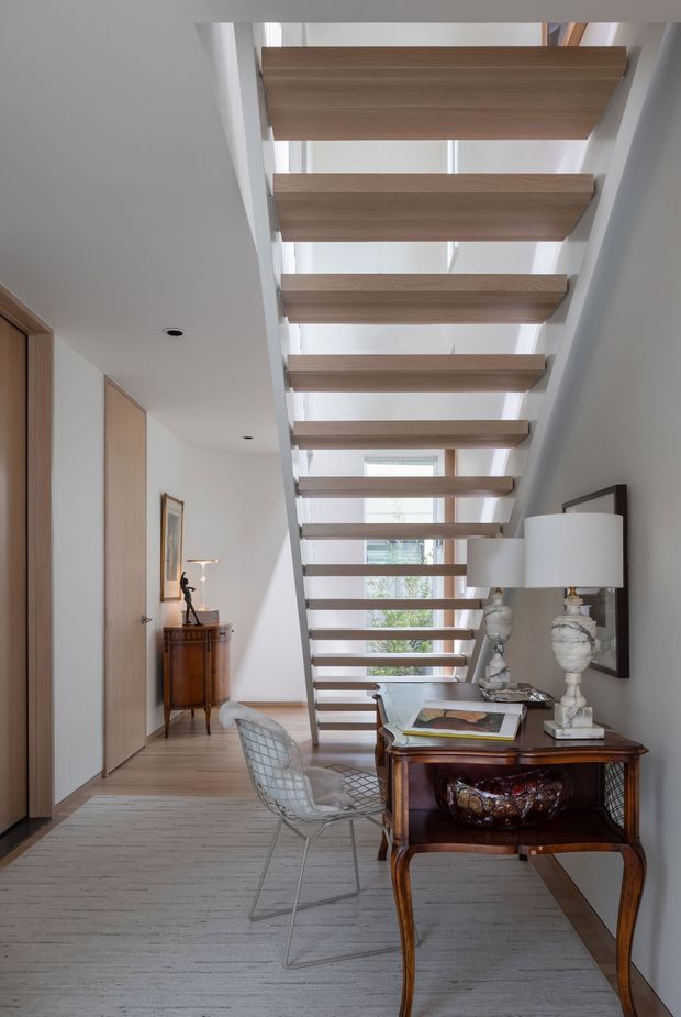 A bright contemporary interior hallway and stairway featuring an open-riser staircase with floating light timber treads and a glazed skylit void above. White walls and ceilings contrast with timber doors and wall panelling. A dark traditional console desk with sculptural lamps and a wire lounge chair sits beneath the stairs, creating a refined transitional mix of modern architecture and classic furnishings.