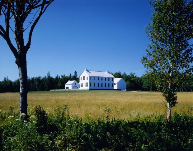 A white multi-storey farmhouse-style building with a symmetrical gabled roof, tall rectangular windows, and attached smaller wings, situated in an expansive rural field. Mature trees and hedges frame the foreground, with a clear blue sky and wooded horizon beyond.