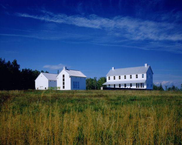Exterior view of two white, traditional-style rural buildings set in an open grassy field. The larger two-storey building has a simple rectangular form, symmetrical rows of windows, a pitched pale metal roof, and dark shutters or openings along the lower level. A smaller adjacent gabled building has matching white walls, a pitched metal roof, and tall narrow windows. The scene is framed by trees and a deep blue sky.