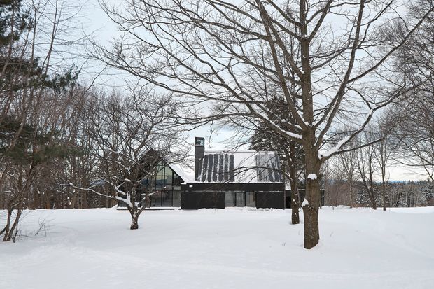 A modern dark-clad house viewed from across a snow-covered yard, surrounded by leafless deciduous trees and woodland. The building has a simple rectilinear form, extensive glazing, and a steeply pitched metal roof with a chimney or service shaft. The scene is a quiet winter exterior with muted natural colours.