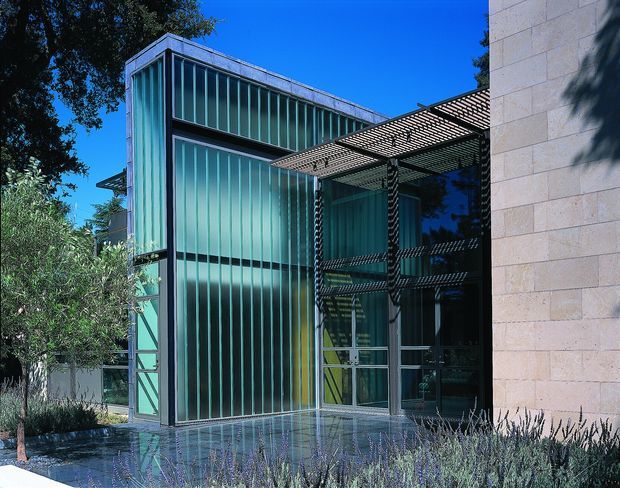 Exterior view of a contemporary building entry or glazed façade, featuring tall turquoise translucent ribbed glazing in dark metal frames, a projecting slatted metal canopy, and a light beige masonry wall. Reflective paving and landscaped planting sit in the foreground beneath a clear blue sky.