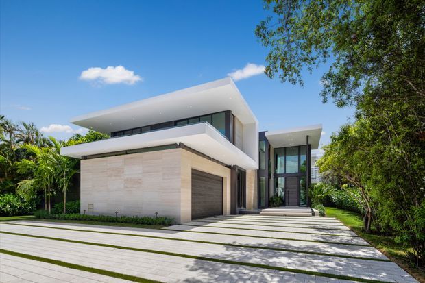 A contemporary two-storey residence viewed from the front, with strong horizontal flat roof planes, white rendered volumes, pale stone-clad walls, extensive dark-framed glazing, and a recessed garage. A broad paved driveway with linear grass joints leads to the entry, surrounded by mature tropical landscaping and trees under a clear blue sky.