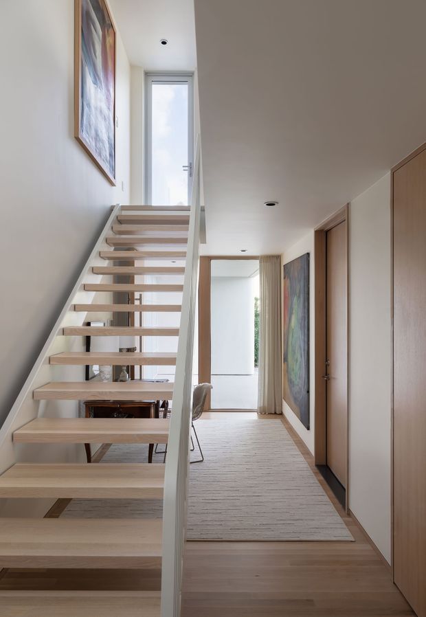 Bright minimalist interior view of a stair hall and landing. An open-riser timber staircase with a slim white balustrade rises toward a narrow glazed door or window, while pale timber flooring and built-in timber cabinetry line the hallway. Recessed ceiling lights, framed artwork, a neutral rug, and a glimpse of a dining area create a clean, airy composition.