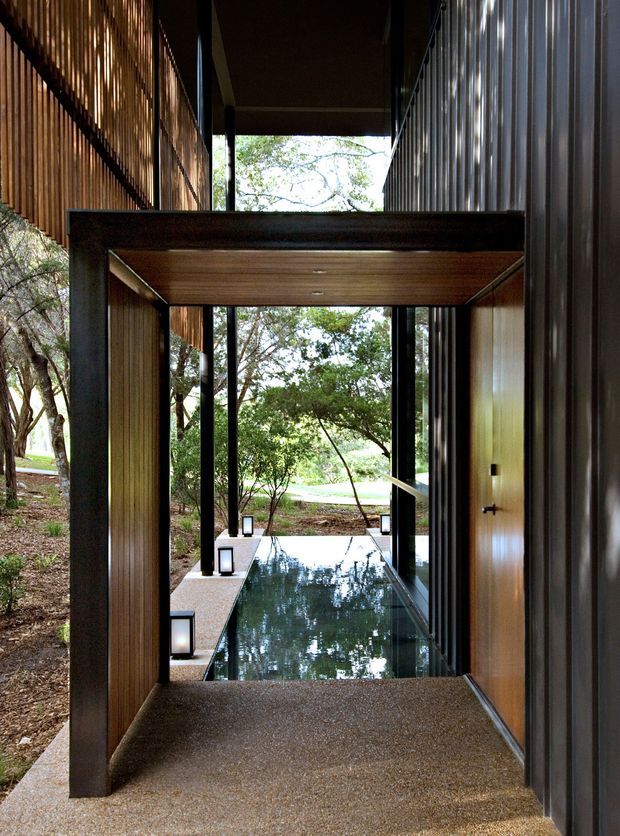 A dramatic covered passage or entryway framed by dark steel posts, vertical timber slat walls, and corrugated metal cladding. A shallow rectangular reflecting pool runs along the centre, bordered by textured paving and small lanterns, while large openings reveal a wooded landscape beyond. The architecture has strong linear geometry, warm timber soffits, and a dark, contemporary material palette.
