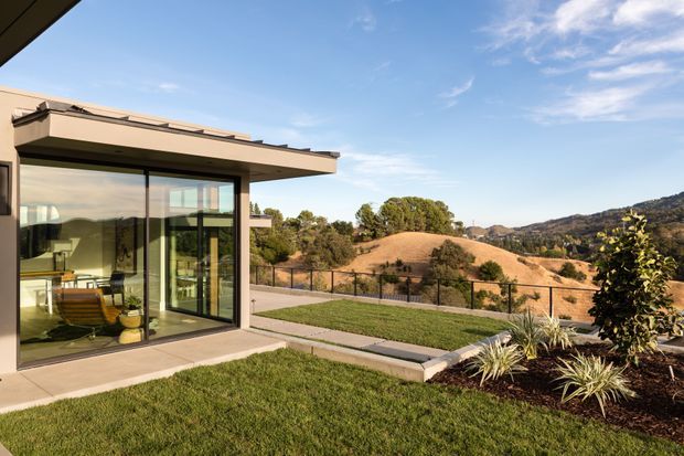 Exterior view of a low-profile contemporary home with broad floor-to-ceiling glazing opening onto a paved terrace and landscaped lawn. The house has a projecting flat roof, slim dark window and door frames, and a minimalist facade. Through the glazing, a bright interior seating and dining area is visible. The garden includes raised concrete-edged planting beds, ornamental grasses, and a wire fence, with dry rolling hills in the background.