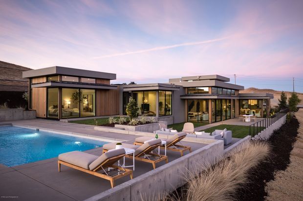 A contemporary single-storey residence with a low, flat-roofed composition and extensive floor-to-ceiling glazing, arranged around a landscaped terrace and swimming pool. The home features dark framed glass walls, warm timber-clad sections, pale rendered or concrete walls, and multiple projecting roof planes. Outdoor loungers and seating occupy the pool terrace, with ornamental grasses and sparse landscaping surrounding the property at dusk.