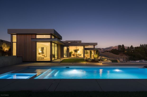 Twilight exterior view of a contemporary multi-wing residence with strong horizontal flat rooflines, extensive floor-to-ceiling glazing, dark timber wall cladding, and illuminated interiors. A large illuminated swimming pool occupies the foreground, with landscaped grounds and distant hills beyond the property.