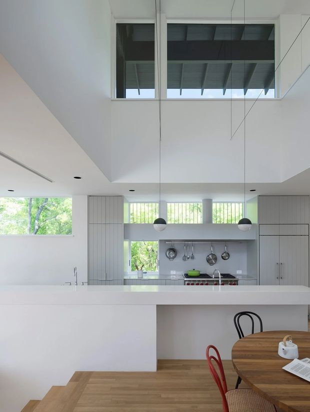Bright contemporary open-plan kitchen and dining area with a large white island, integrated pale cabinetry, stainless-steel cooking appliances, pendant lights, and warm timber flooring. The space features a dramatic double-height void with high clerestory glazing and a visible roof structure, while a round timber dining table occupies the foreground.