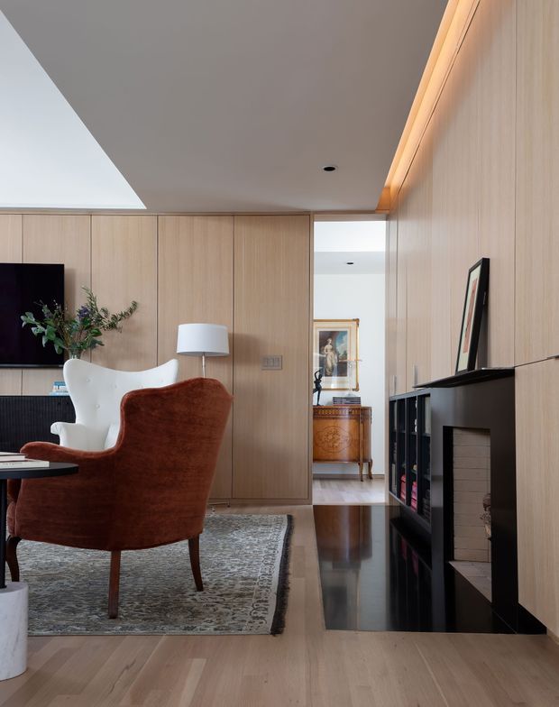 A refined contemporary living room with full-height light timber wall panelling and matching built-in cabinetry, a black-framed fireplace and shelving, polished dark hearth surround, timber flooring, and a patterned area rug. The room includes a sculptural rust-brown and white lounge chair, side table, lamp, television, and artwork visible through the opening beyond. Warm concealed lighting highlights the timber joinery, while a large angled ceiling opening brings in natural light.