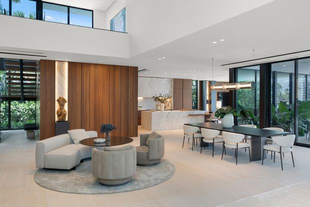 A spacious, open-plan contemporary interior combining a living room, dining area and kitchen beneath a high ceiling with a partial upper-level void. The living zone has curved neutral sofas and ottomans on a round rug, while the dining area features a dark rectangular table with upholstered chairs. A marble-look kitchen island, extensive timber wall panelling, black-framed glazing, indoor planting and sculptural pendant lighting create a refined, warm-modern atmosphere.