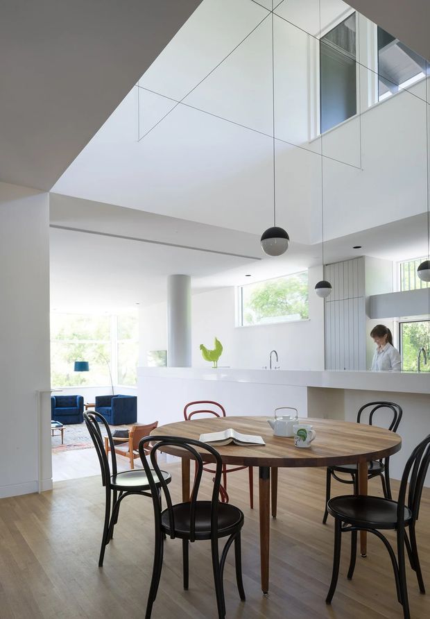 Bright, minimalist open-plan dining and kitchen interior with a large round timber dining table surrounded by black and burgundy bentwood-style chairs. White walls, built-in cabinetry, a peninsula kitchen, pendant lights, expansive windows, and a double-height void with reflective wall panels create an airy contemporary space. A living area is visible beyond the dining zone.
