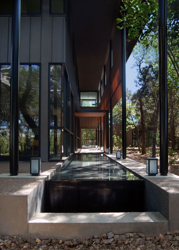 A dramatic modern architectural walkway beside a reflecting water channel, framed by tall black structural columns, floor-to-ceiling glazing, dark cladding, and warm timber-lined walls. The covered passage extends through a wooded landscape, with strong reflections and rhythmic lantern-like fixtures along the edges.