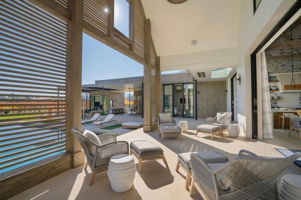 A bright, spacious covered outdoor living terrace beside a pool, furnished with woven lounge chairs, ottomans, and white ceramic side tables. Timber louvred screens and posts frame the space, while large sliding glass doors open into an adjacent interior living or dining area. The terrace has a pale tiled floor, white ceiling, strong indoor-outdoor connection, and views toward a modern pool courtyard.
