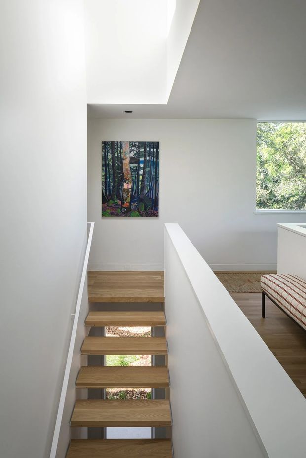 Bright minimalist interior view looking down a stairway with open-riser timber treads, white walls, and a simple white balustrade. Daylight enters through a large skylight or roof opening above and a window beside the upper landing. A colourful forest-themed artwork hangs on the wall, while a patterned upholstered bench is partially visible in the adjacent upper-level space.